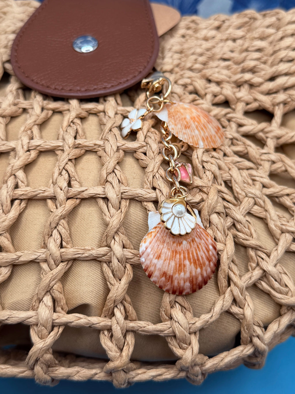 Chocolate seashell bag charm hanging on woven straw crossbody bag with shell wallpaper background