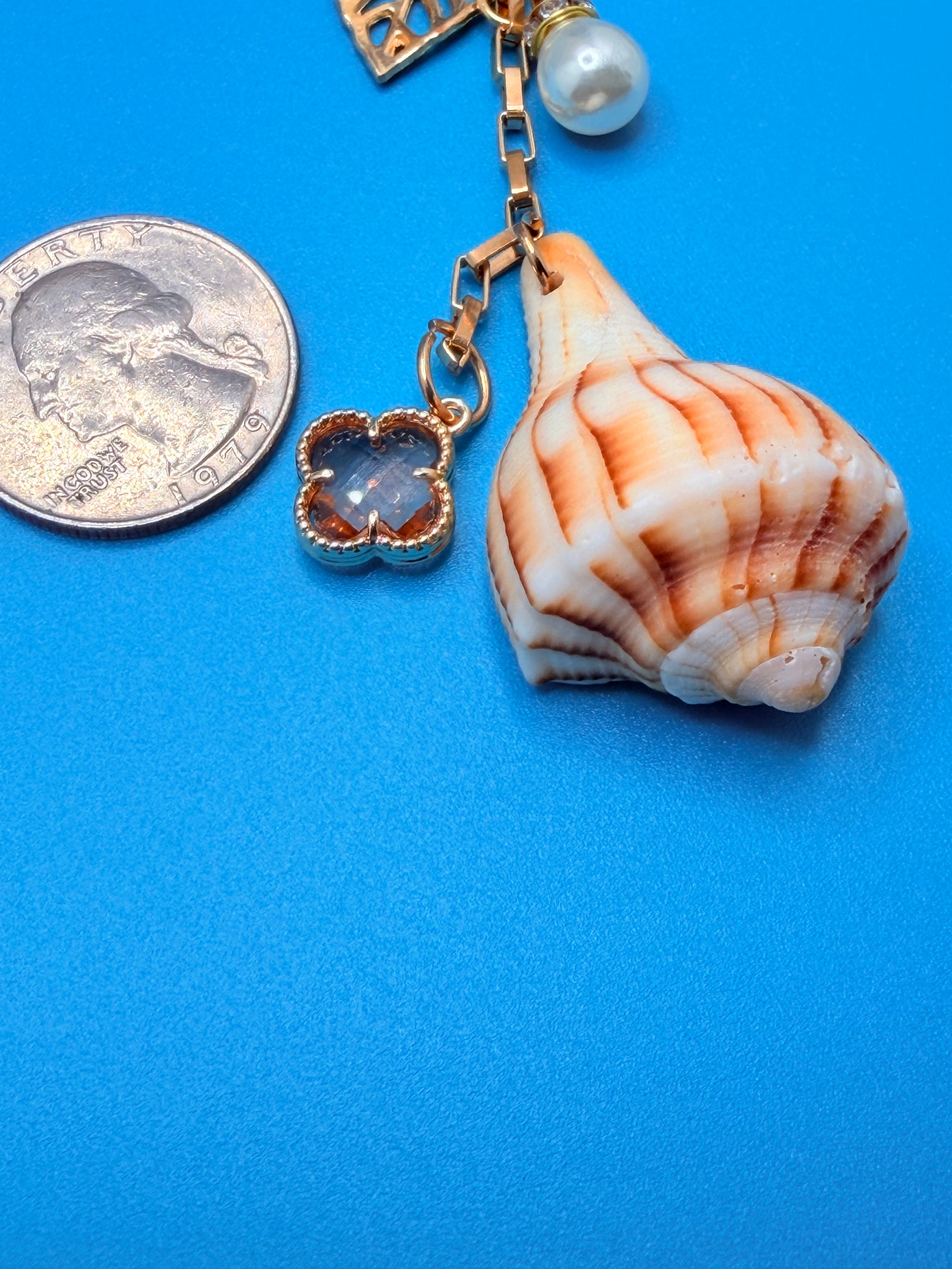 Celestial Shores lightning whelk shell bag charm with quarter for scale on blue background showing amber clover charm