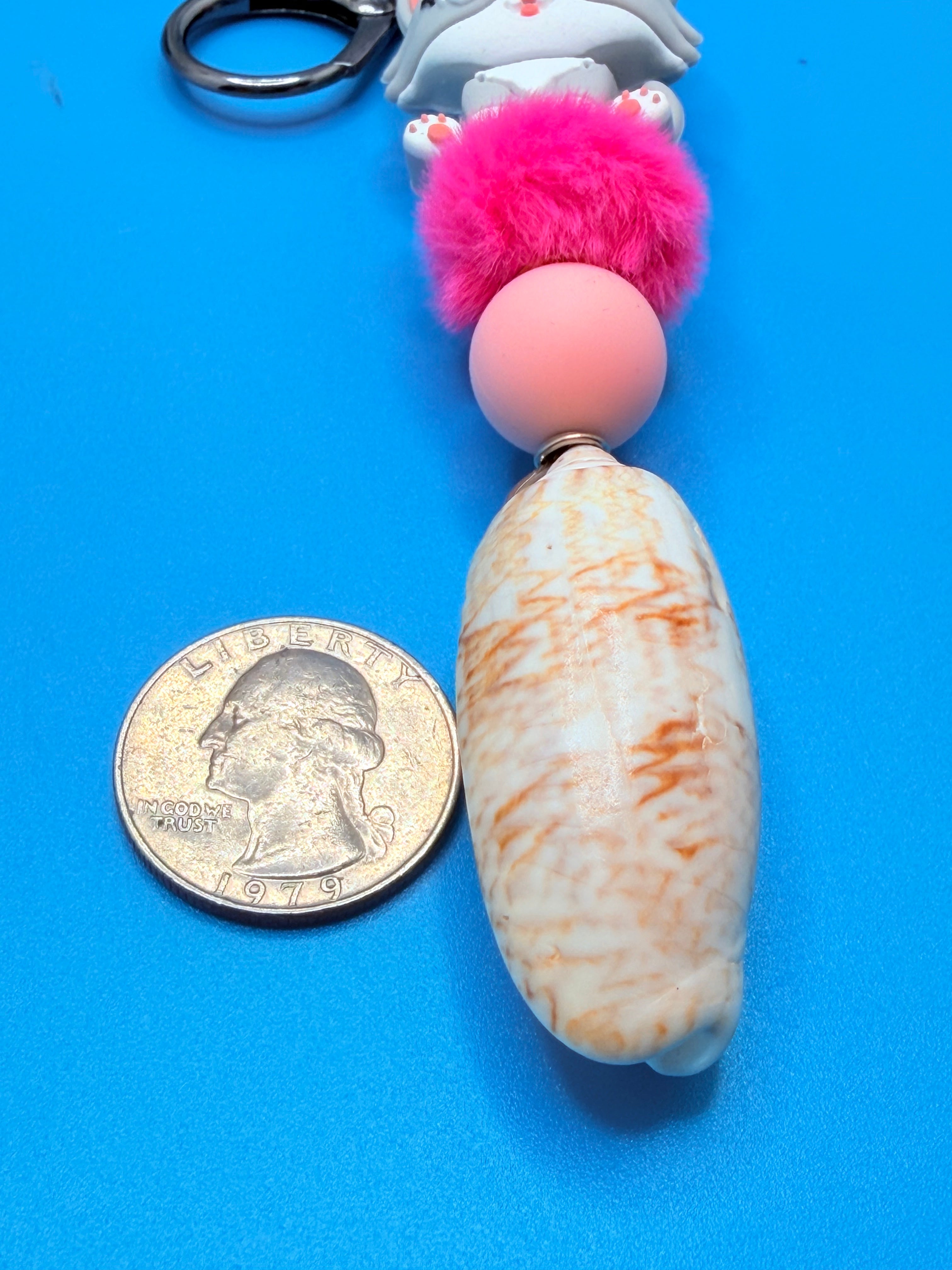 Pink Beret Cat lettered olive shell keychain with quarter for scale on blue background showing lettered olive shell close-up