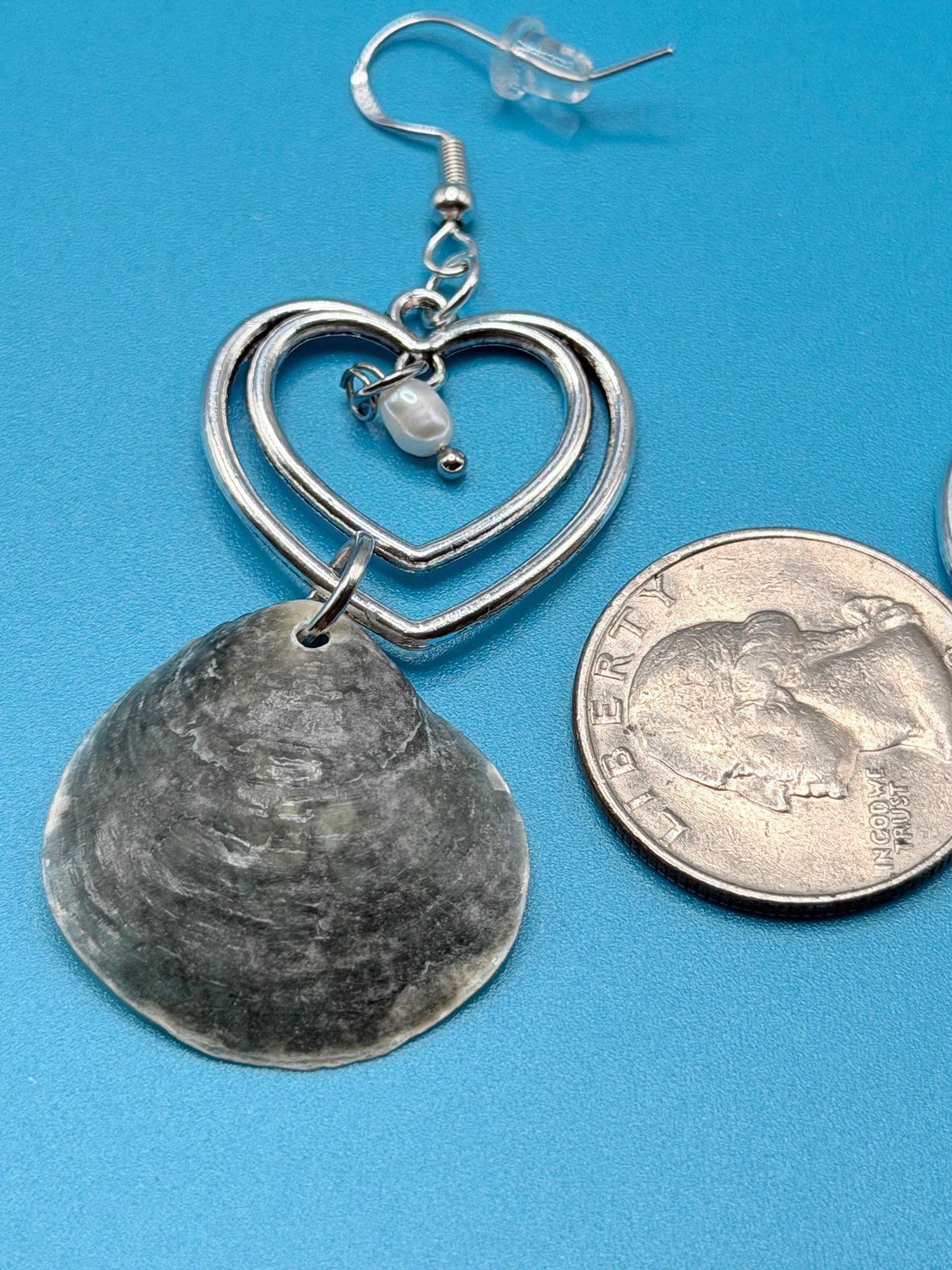 Close-up of single gray jingle shell double heart earring with pearl drop and dime for scale