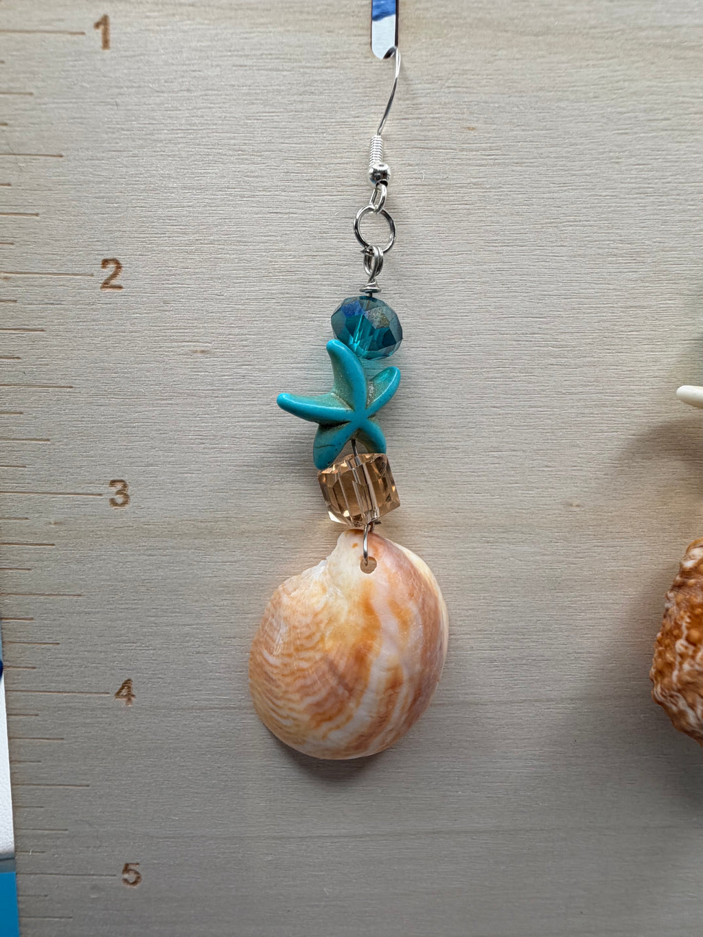 Close-up of slipper shell earring with white starfish charm and teal crystal on ruler