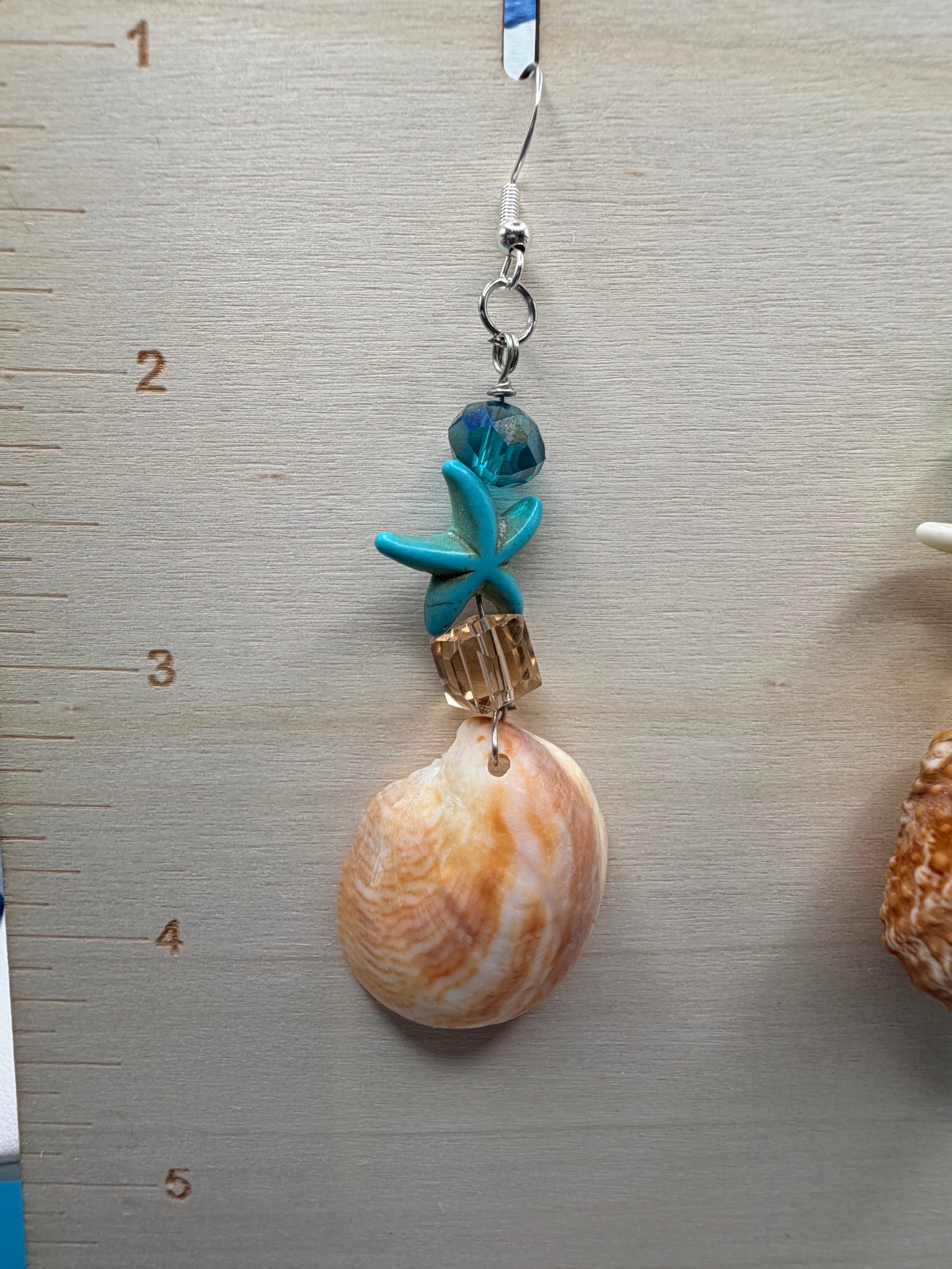 Close-up of slipper shell earring with white starfish charm and teal crystal on ruler