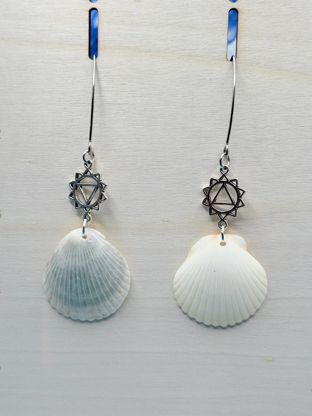 White and gray scallop shell chakra earrings on blue background with quarter for scale
