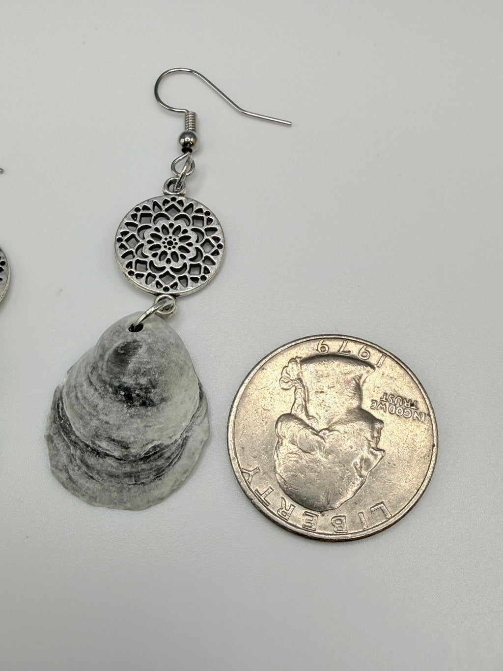 Jingle shell earrings with silver mandala charm next to quarter on coastal background