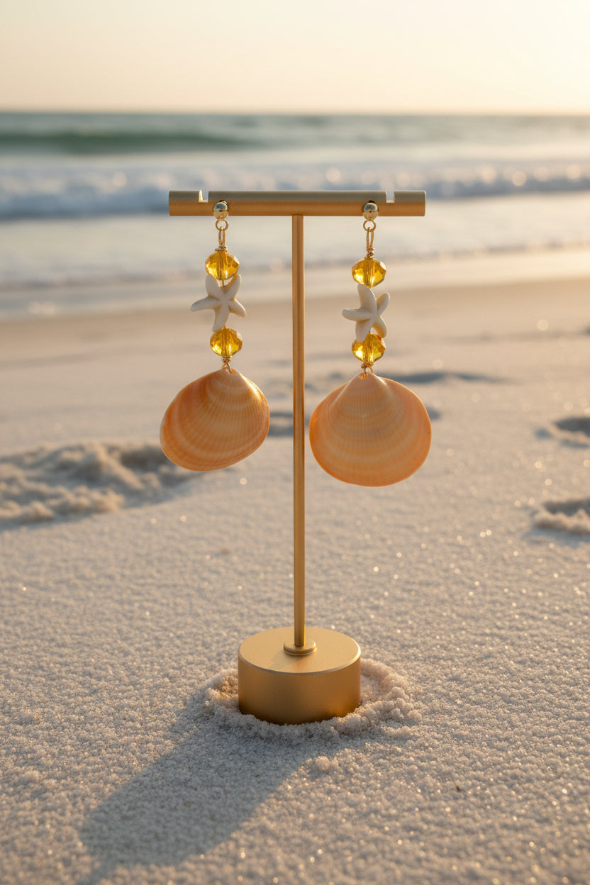 Golden cockle shell and starfish dangle earrings on gold T-bar stand with blue scallop shell backdrop close-up