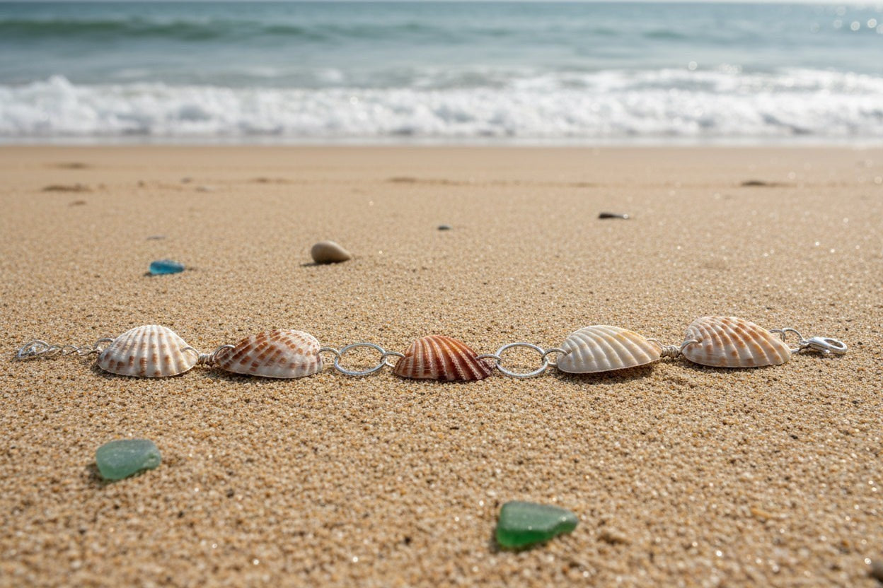 Shoreline Keepsakes cardita shell bracelet with silver links resting on sandy beach with ocean waves in background.