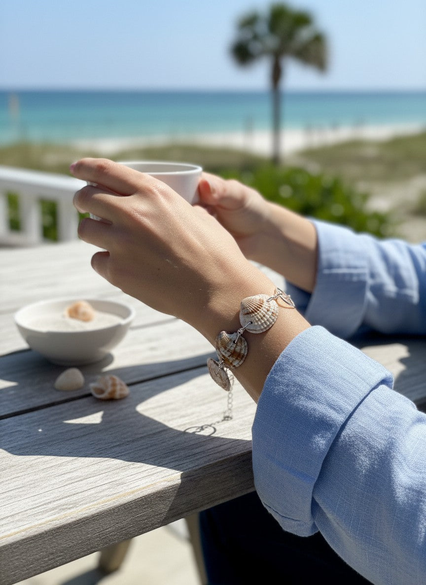Shoreline Keepsakes seashell link bracelet with silver-tone hardware worn on a wrist at a beachside table.