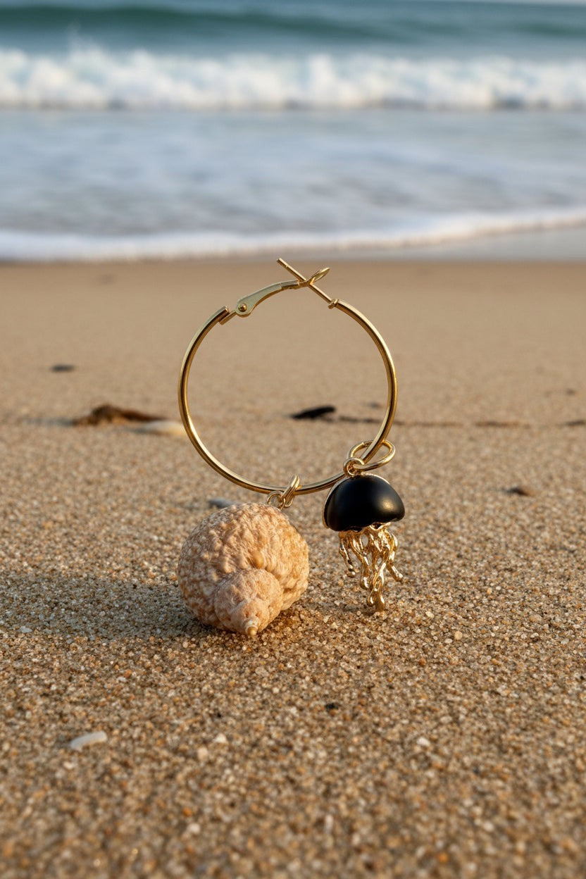 Shoreline Keepsakes gold hoop earring with a turban shell, matte black bead, and gold jellyfish charm on sandy beach